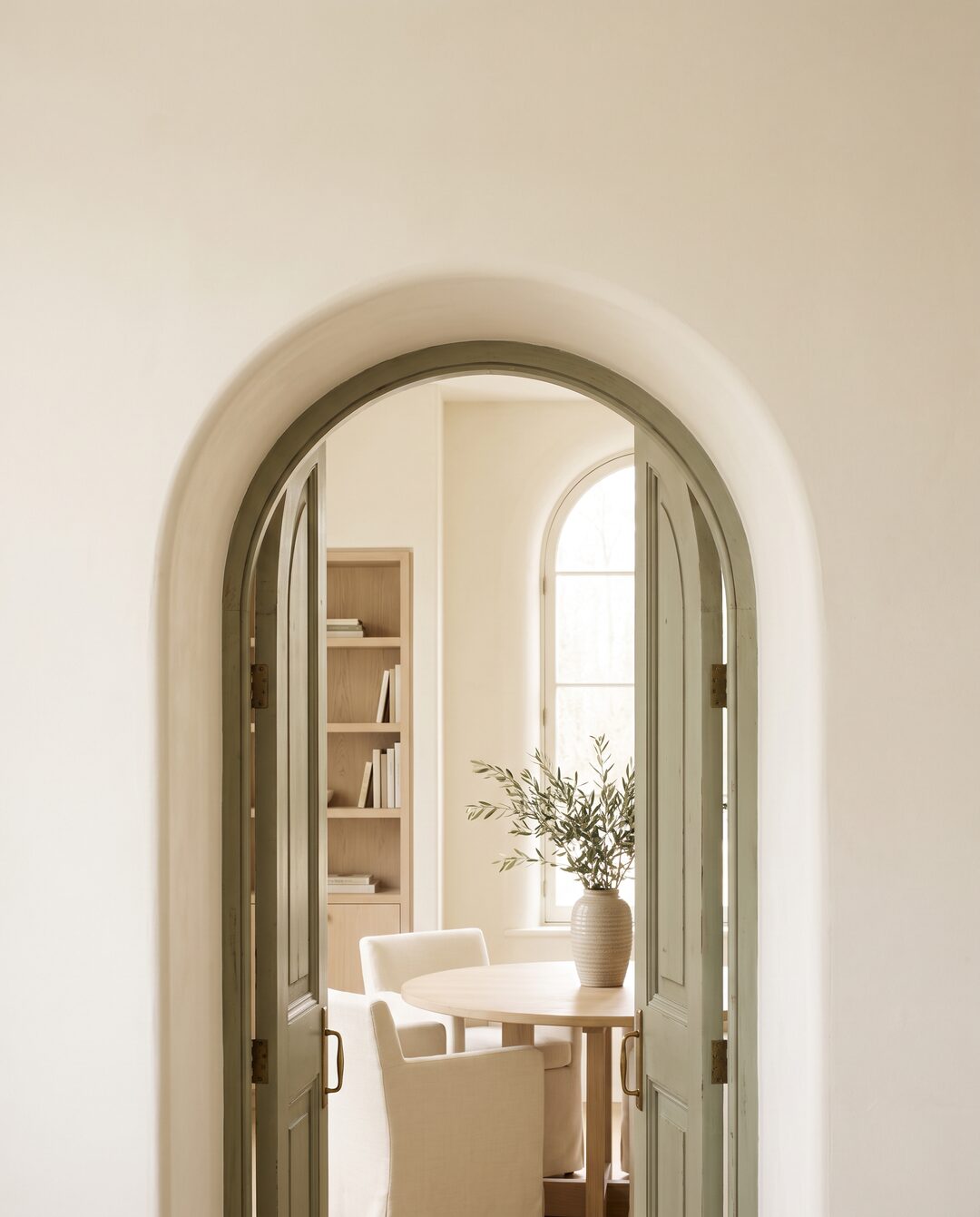 Ein Rundbogen aus cremefarbenem Putz, darin geöffnete salbeigrüne Flügeltüren; dahinter ein heller Raum mit Bücherregal, rundem Tisch und einem Olivenzweig in einer Vase.